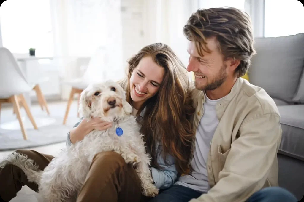 A happy couple sitting on the floor with their fluffy white dog in a bright, modern living room.